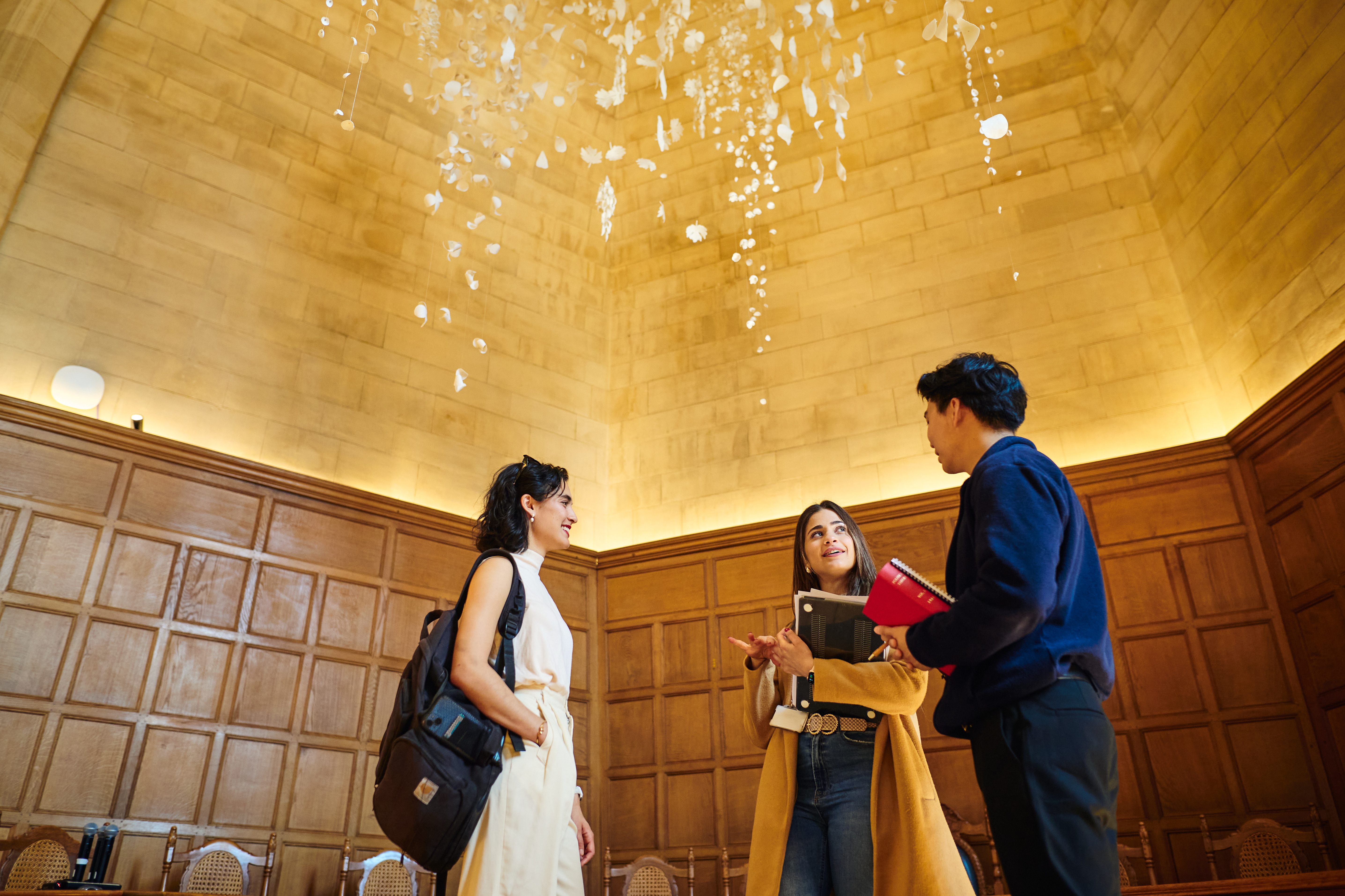 Three Scholars engaged in a lively discussion, standing under the Floating Garden sculpture with books and papers in their arms.