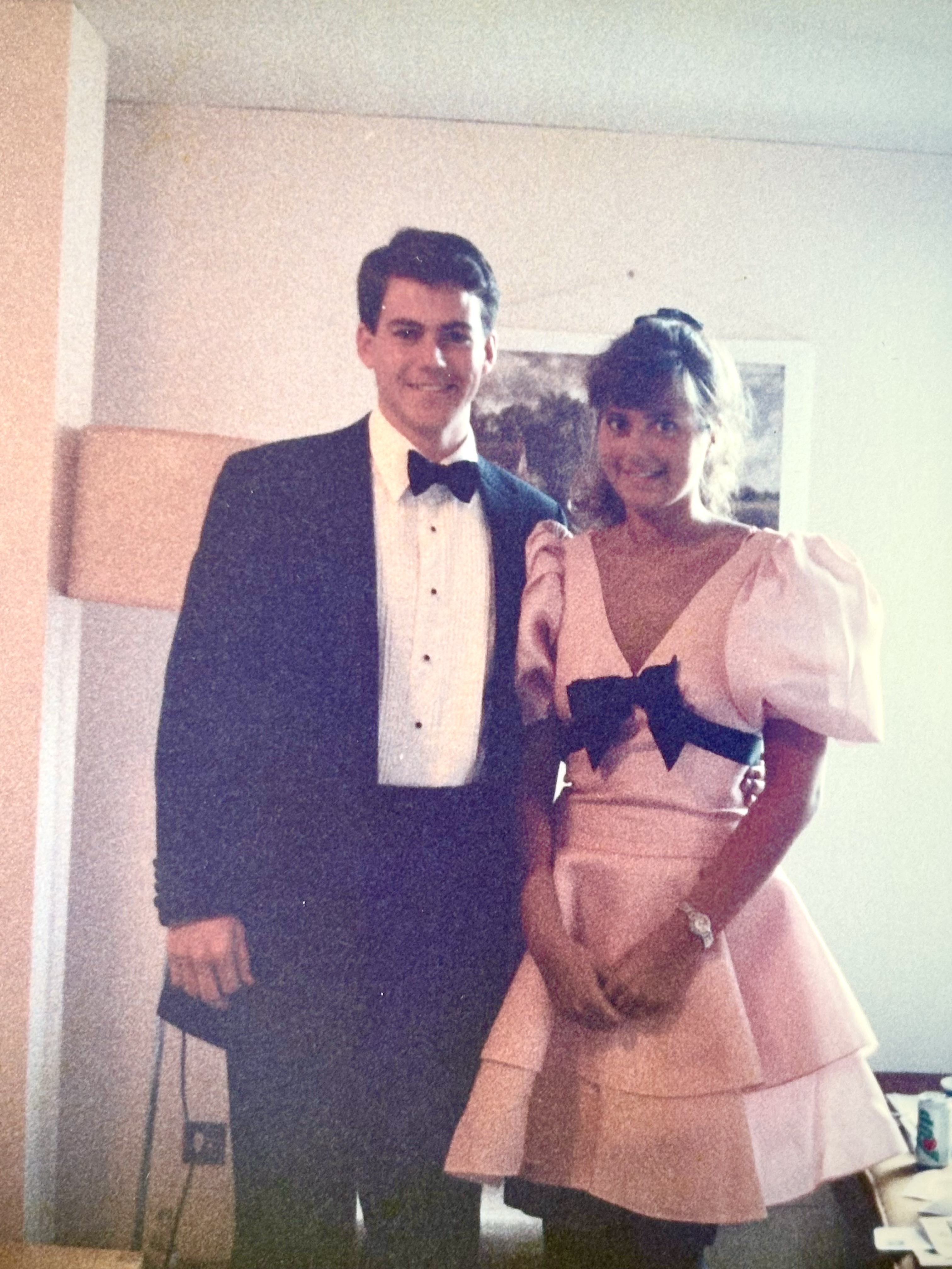 David stood to the left in a suit and bow tie, and Tamara to his right wearing a short pink frilly dress and ponytail. Both are smiling and appear to be stood in a bedroom, with a lampshade and framed image in the background.