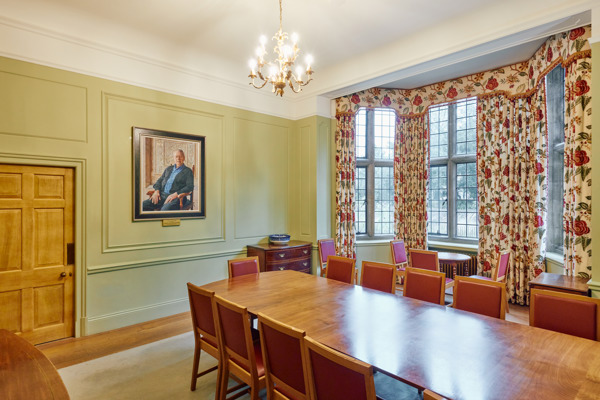 A whole room shot of the Atlantic Room in Rhodes House. This angle looks diagonally down from the head of a long wooden table. On the left wall is a large wooden door and a portrait of a gentleman sat in a chair. In the wall to the right of this is a large bay window framed by floral curtains.