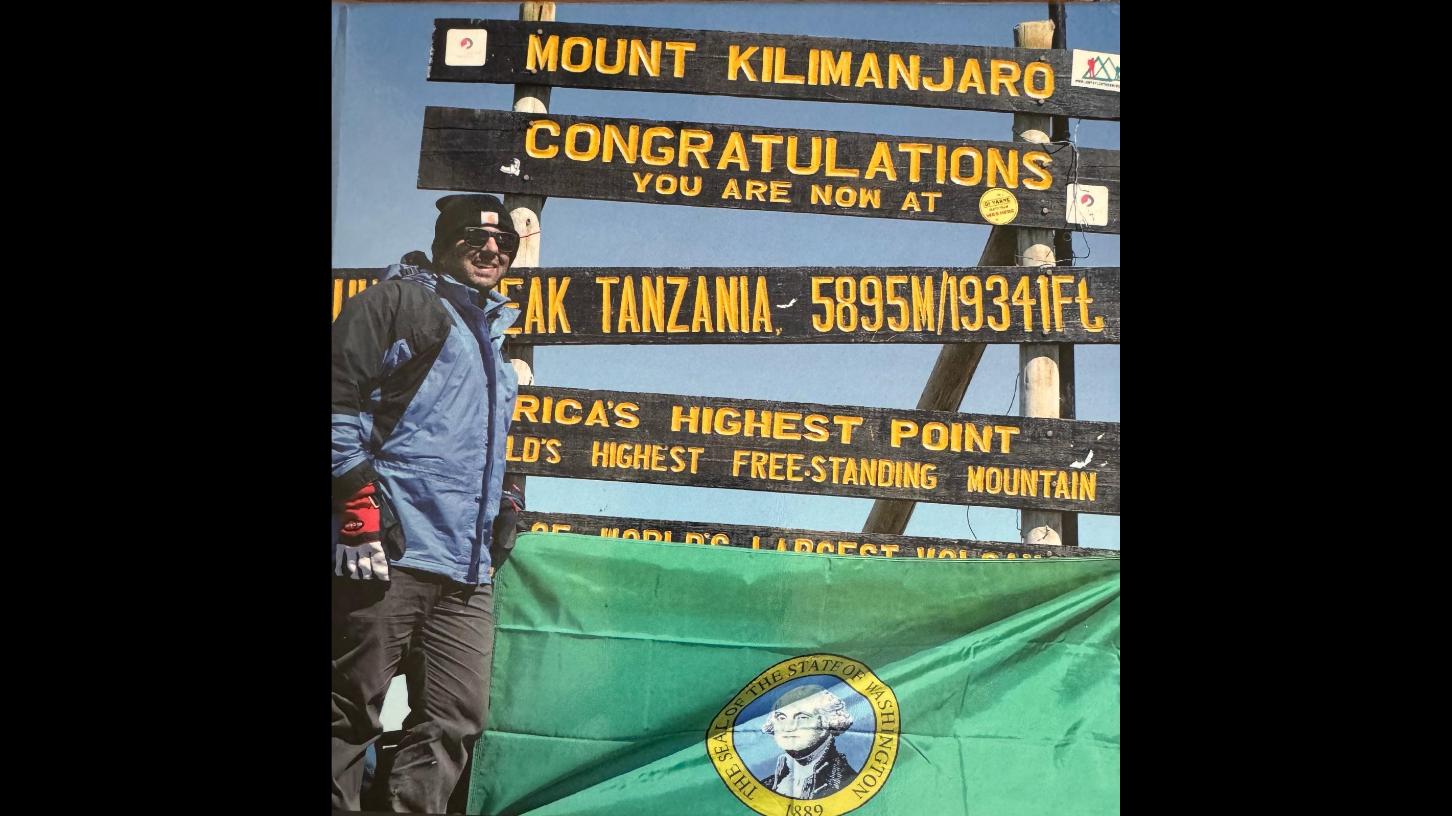 The photo shows Cyrus on the peak of Mt Kilimanjaro with a green flag bearing the seal of the state of Washington 
