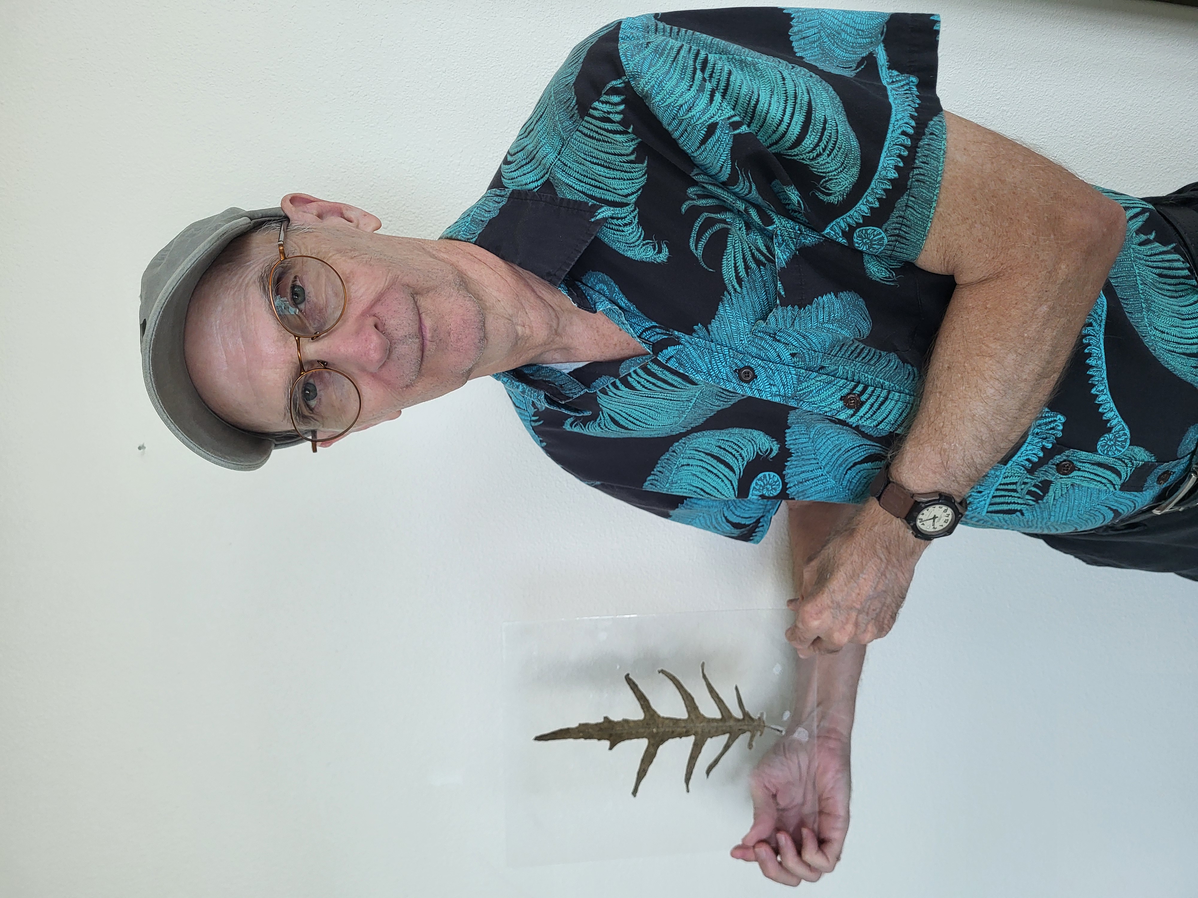 Roger is holding a clear case with the a leaf displayed within it. The leaf is green, long and has three stems coming out either side.