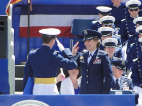 Michelle saluting to another colleague. She is graduating, dressed in uniform. There is a crowd of soldiers sat on tiered seating behind her.