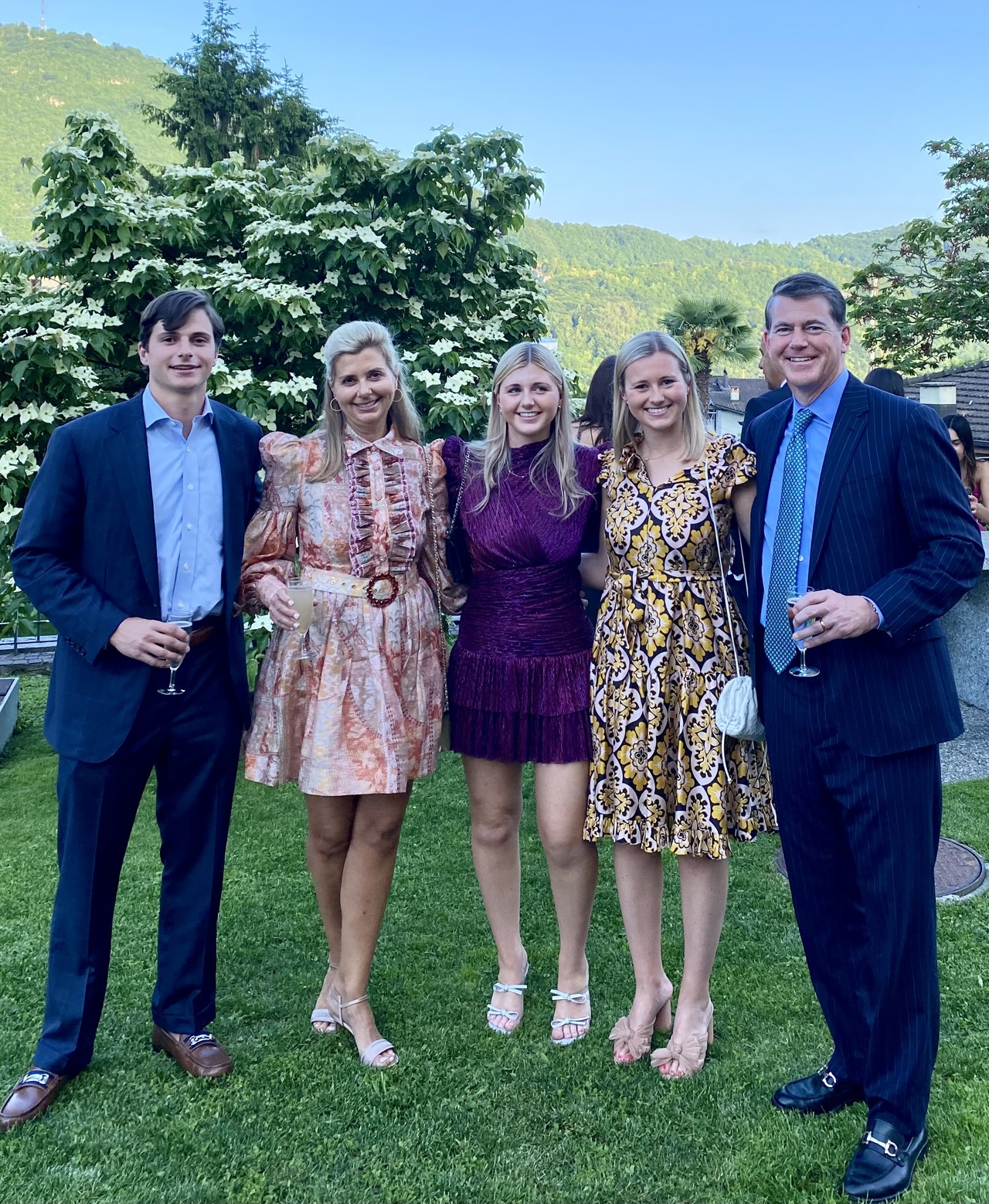 A group of 5 adults stood in dresses and suits. They appear to be in a garden, with hills seen behind them. All are smiling and posing for the camera.