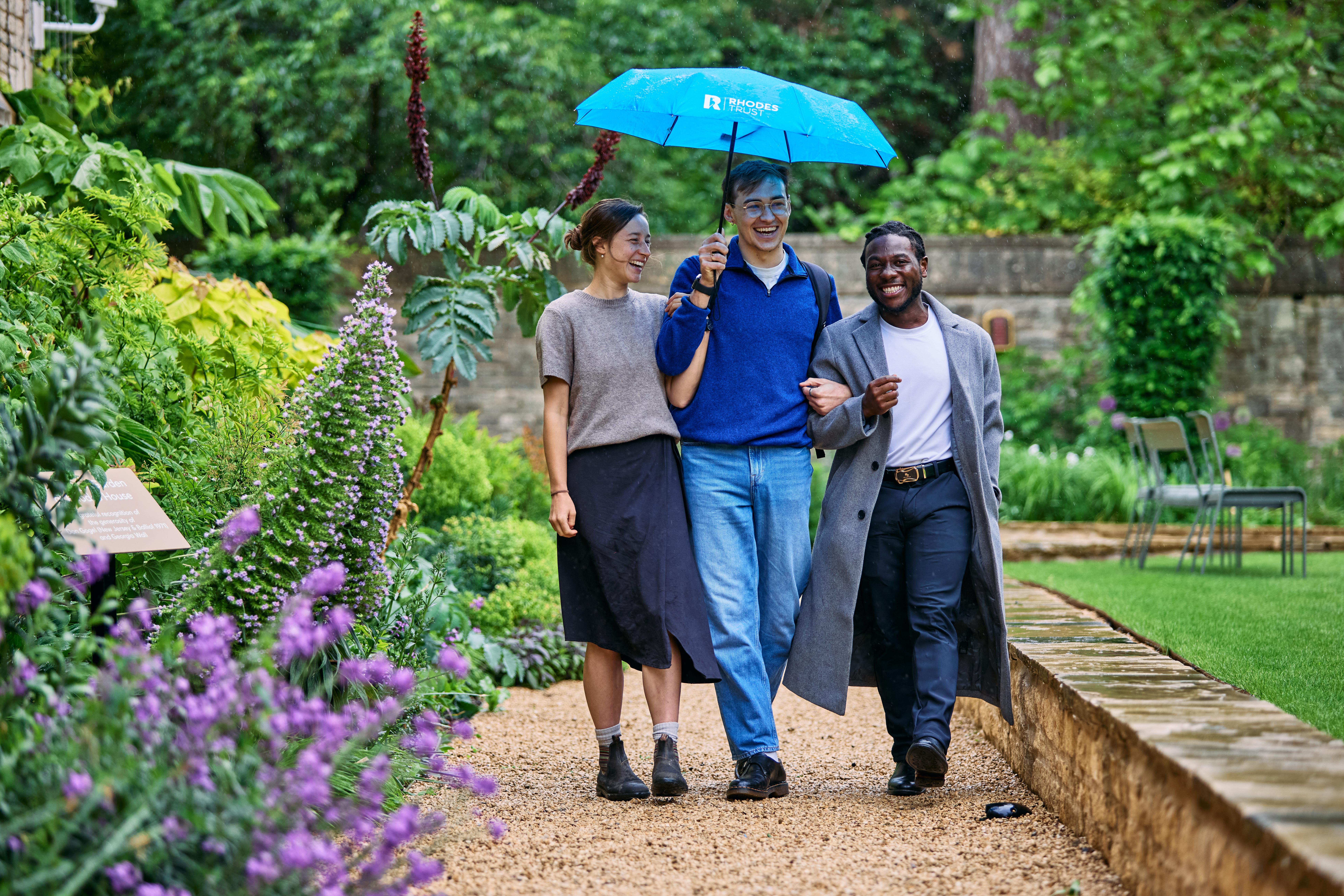 Three Scholars walking arm-in-arm on a garden path at Rhodes House under a shared blue umbrella featuring the Rhodes Trust logo.