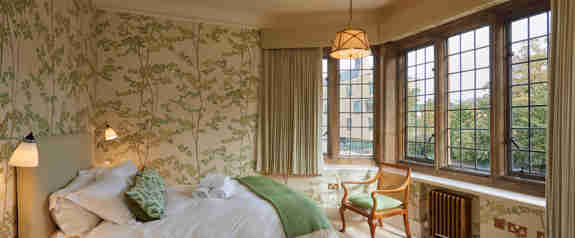 A Bedroom With Green Floral Wallpaper And Green Coloured Soft Furnishings Facing The Bed Are Large Old Fashioned Windows That Allow Light To Flood In
