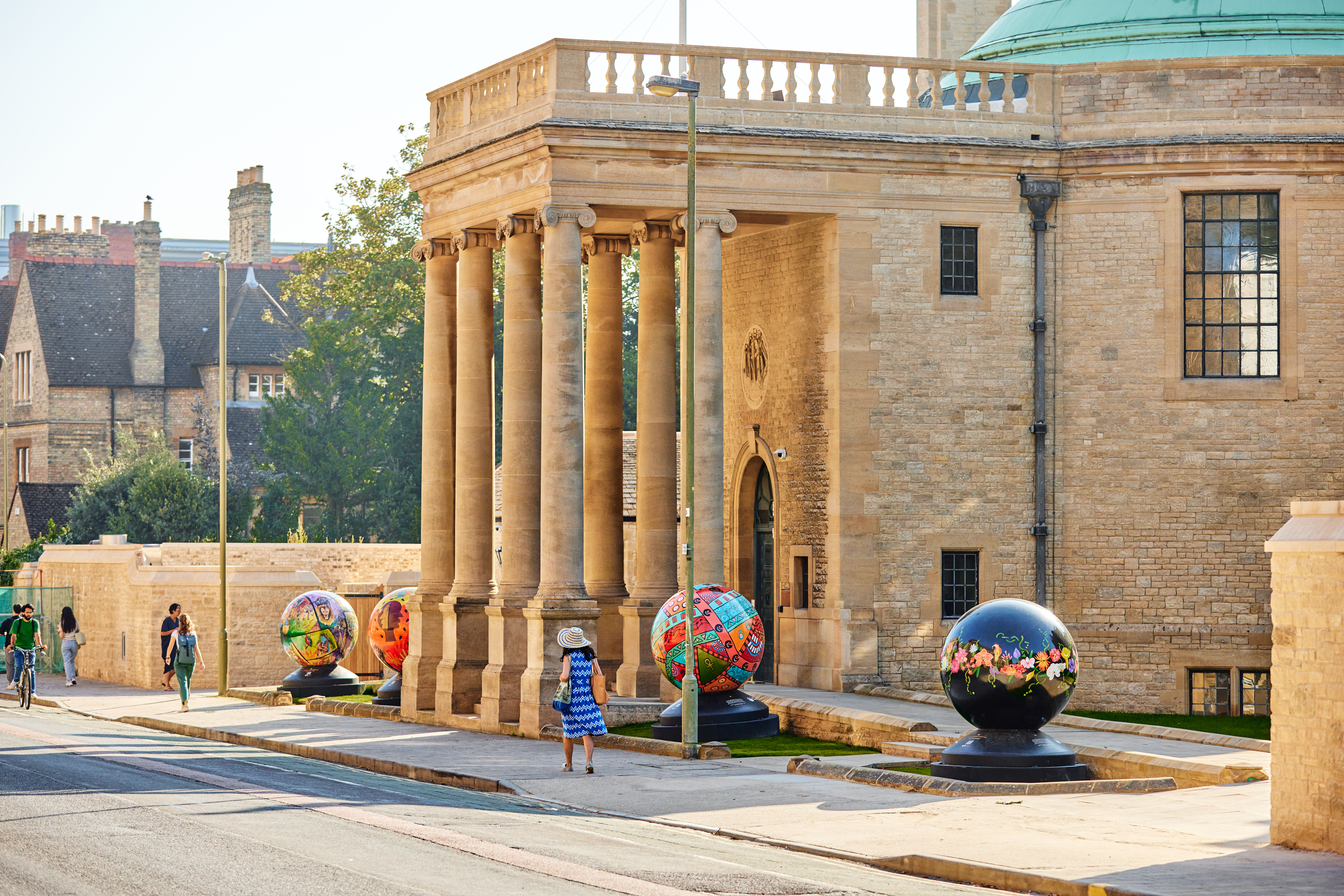 The sun shines brightly on the front of Rhodes House which has four brightly coloured globes by the World Reimagined outside.