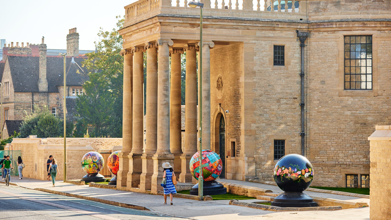 The sun shines brightly on the front of Rhodes House which has four brightly coloured globes by the World Reimagined outside.