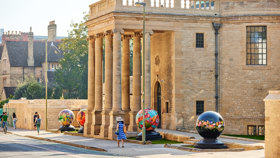 The sun shines brightly on the front of Rhodes House which has four brightly coloured globes by the World Reimagined outside.