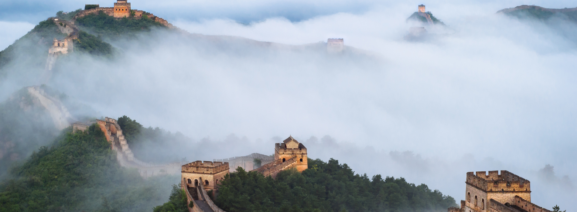 An image of the Great Wall of China from above. There are low clouds over the trees and structures.