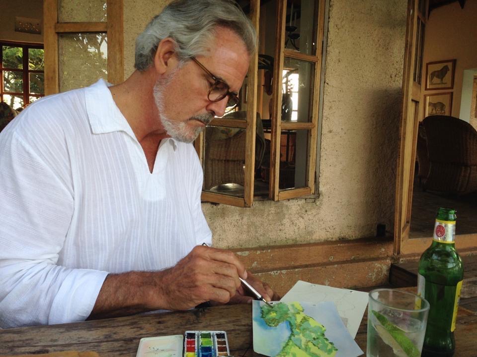 Ken is sat at a wooden table painting a watercolour green landscape.