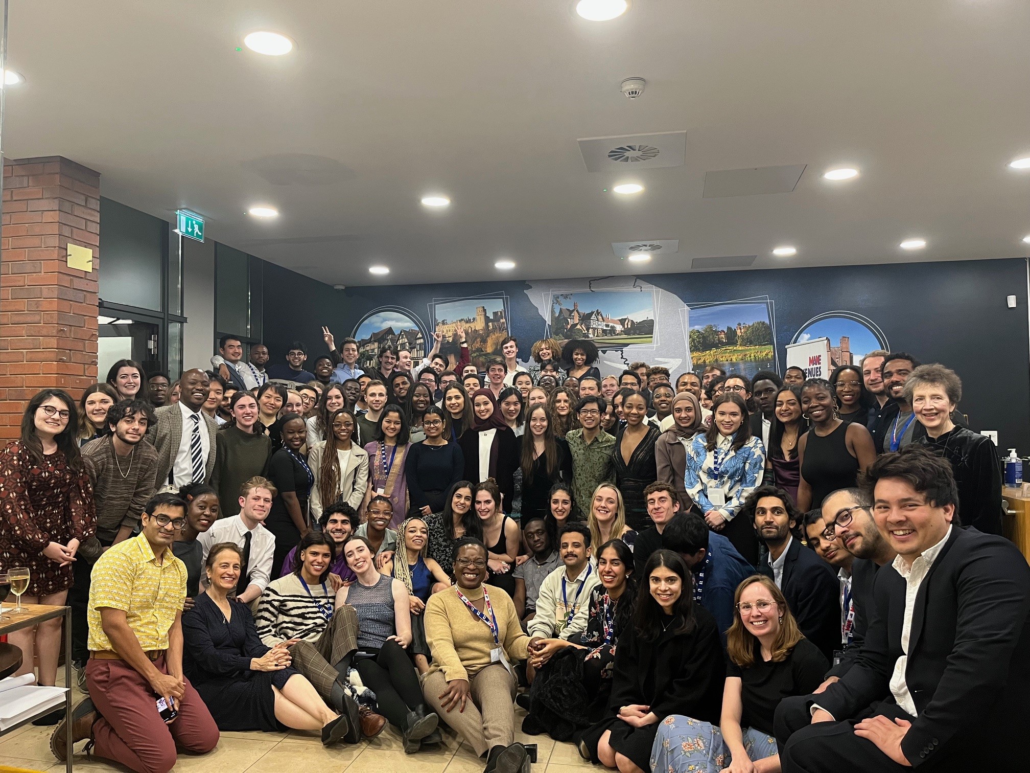 A group photo of around 50 Scholars grouped together posing for a photo. All are smiling and crouching around, Elizabeth is sat on the floor in the bottom left corner, surrounded by Scholars.