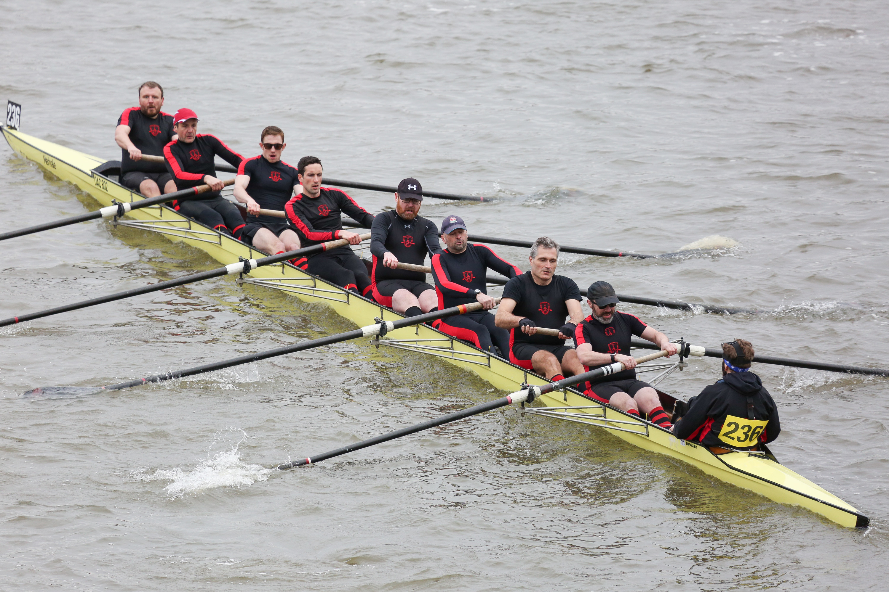 Photograph of a yellow rowing boat with 8 men seen rowing facing the coach.