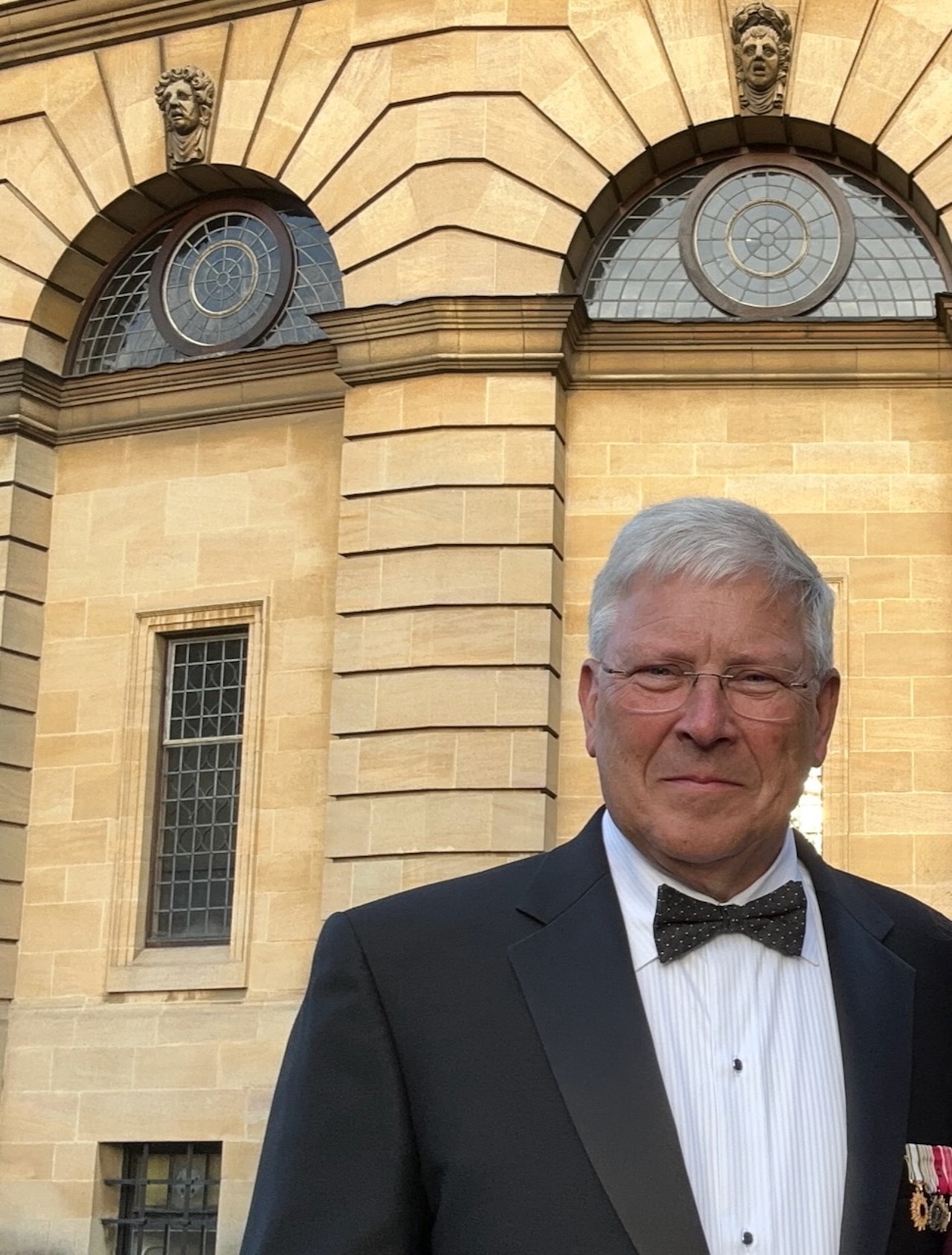 A recent photo of Chris stood in Oxford before the Bodelian wearing a black and white suit with bow tie.