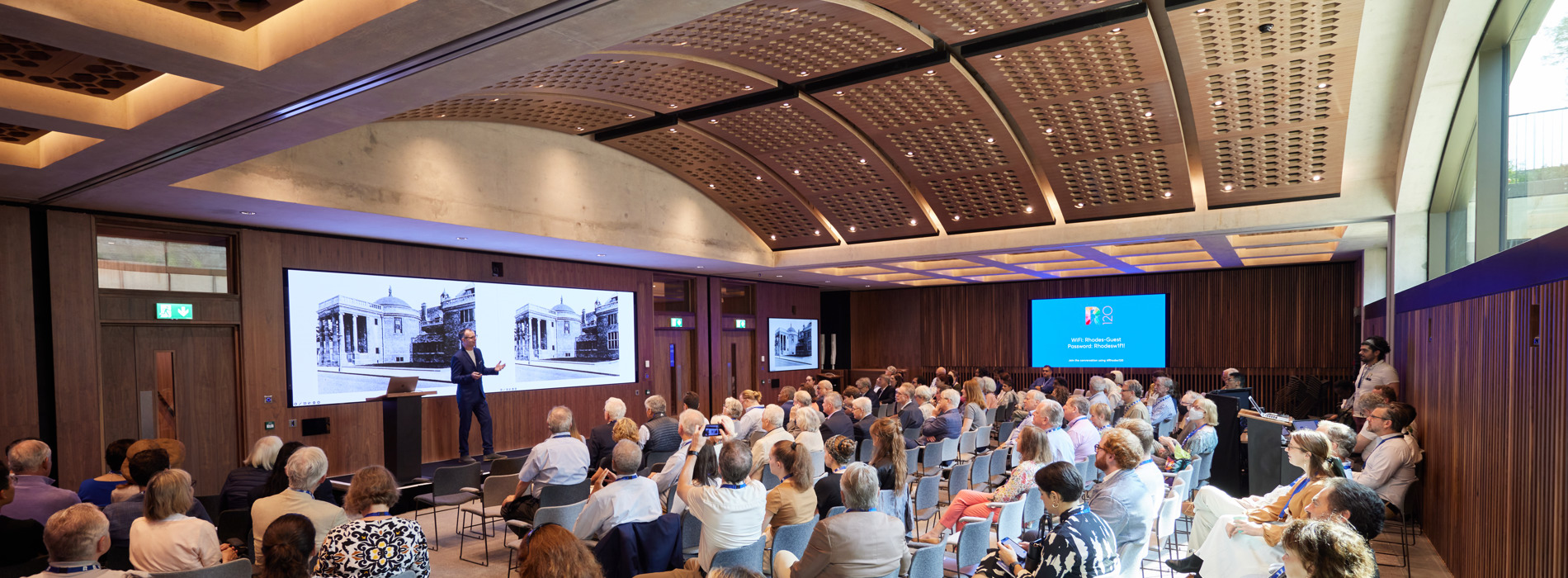 Conference in the Rhodes House Conference Hall with presenter standing in front of the main screen and delegates facing the stage.
