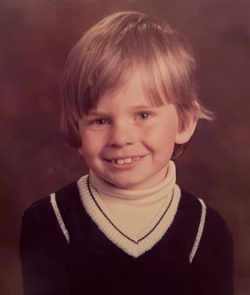 A sepia-tinted picture of Mark as a young boy, in a black sweater, smiling at the camera