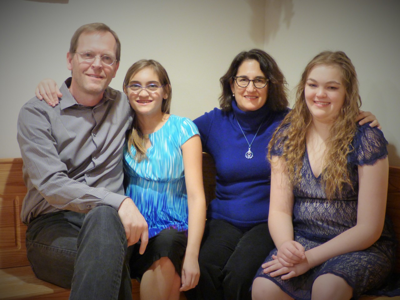 Family of four sat smiling on a wooden bench. Eric to the left, daughter to his right, then his wife, and second daughter on the far right.