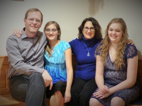 Family of four sat smiling on a wooden bench. Eric to the left, daughter to his right, then his wife, and second daughter on the far right.