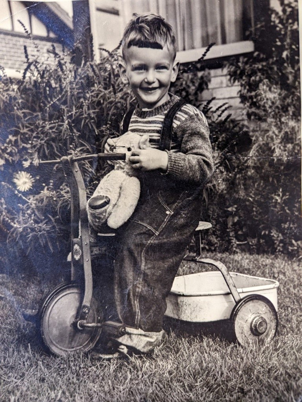 a black-and-white picture showing a young Laurance on a tricycle with a stuffed toy in his hands. Laurance is smiling at the camera, and he is probably in the backyard.