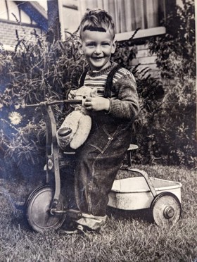 a black-and-white picture showing a young Laurance on a tricycle with a stuffed toy in his hands. Laurance is smiling at the camera, and he is probably in the backyard.