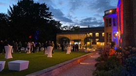 Drinks reception at night on the West Lawn with the Glass Pavilion in the background, poseur tables on the lawn, and red and blue uplighters on the outside walls
