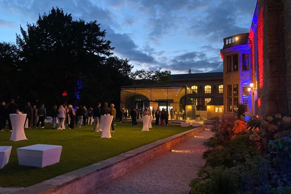 Drinks reception at night on the West Lawn with the Glass Pavilion in the background, poseur tables on the lawn, and red and blue uplighters on the outside walls