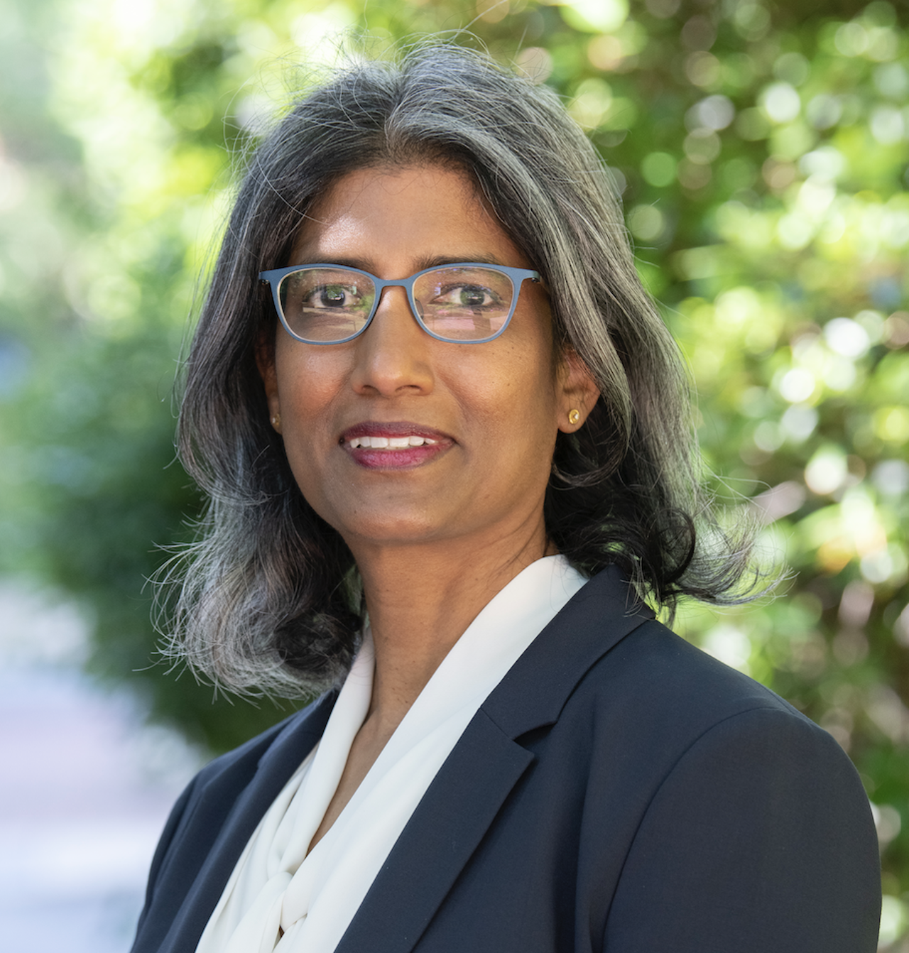 Portrait photo of Ruanne in a black and white suit, wearing glasses, trees behind her.