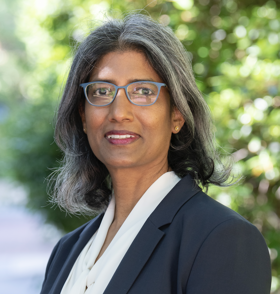 Portrait photo of Ruanne in a black and white suit, wearing glasses, trees behind her.