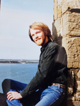 Eric sat on a stone structure wearing blue jeans and a shirt. He is looking into the distance, and a bright blue sea and sky is seen behind him.