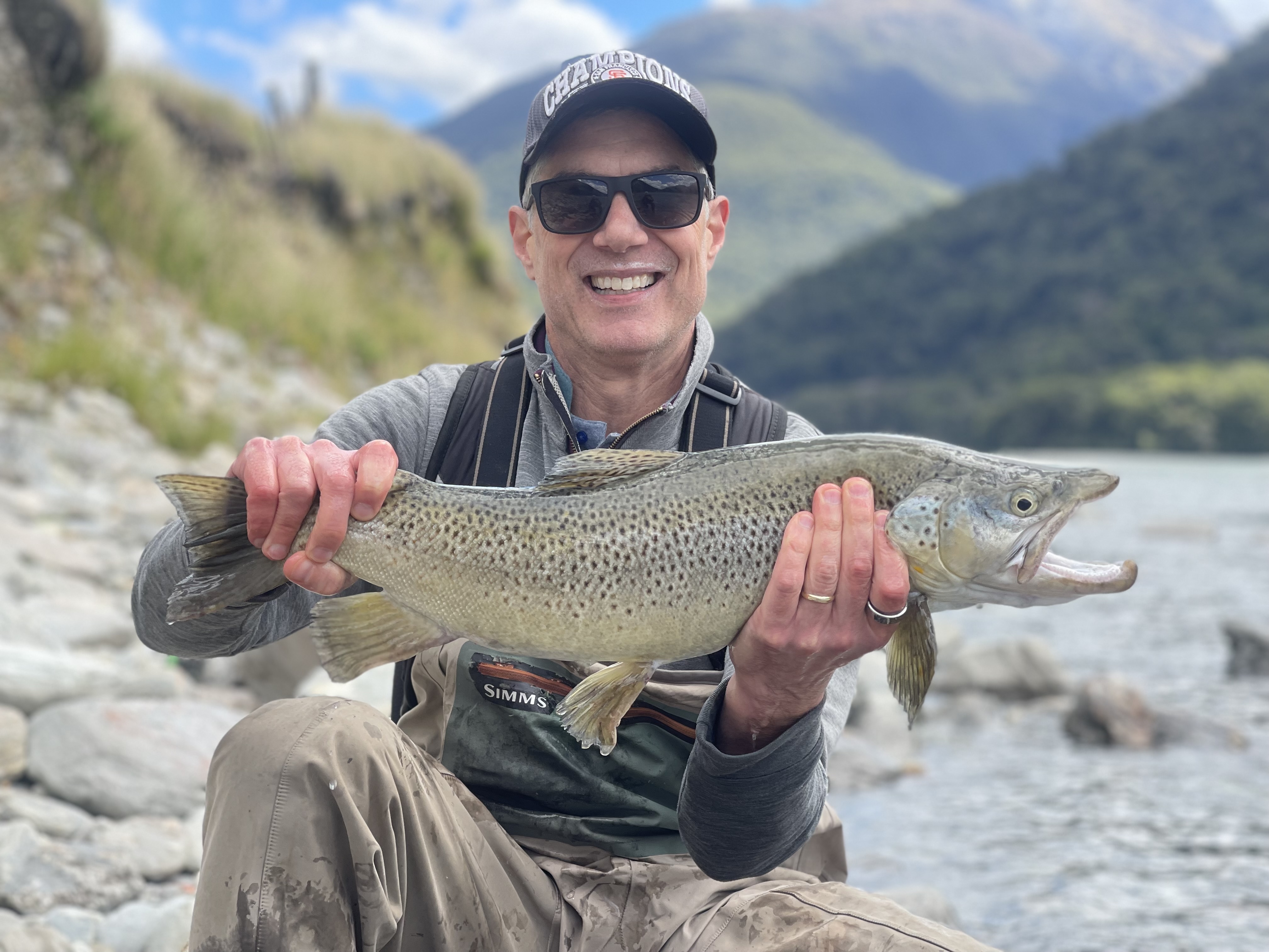 Paul leaning down next to a river holding a fish and smiling. He is wearing a cap and sunglasses, the view behind him is of green mountains and a blue sky.