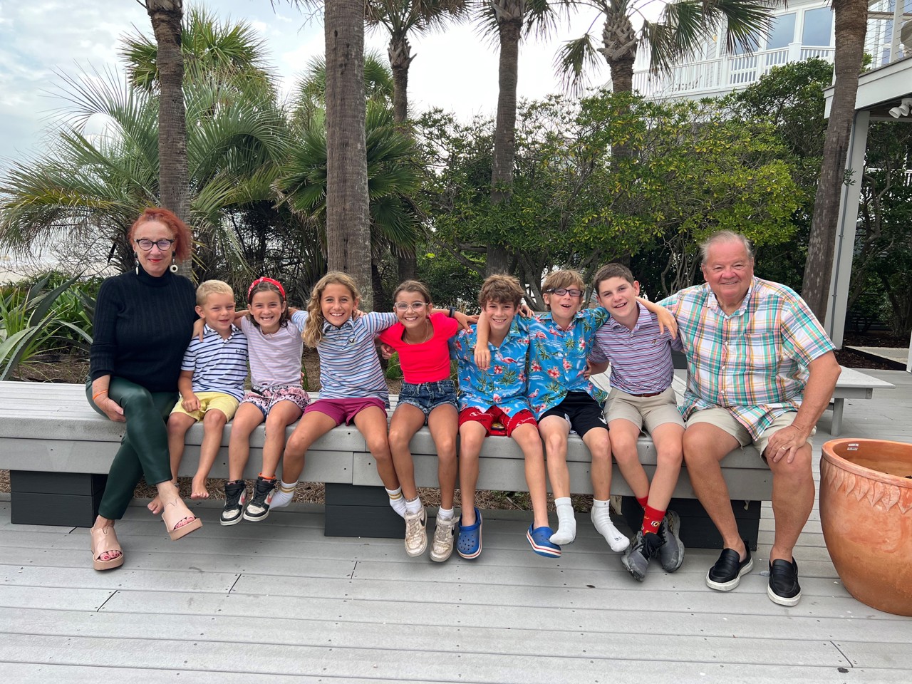 Nick and Marla either side of their 7 grandchildren all sat on a bench with palm trees behind them.