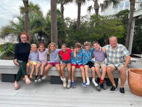 Nick and Marla either side of their 7 grandchildren all sat on a bench with palm trees behind them.