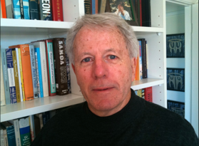 A portrait of Peter. He's standing in front of a wall of books. He is in a black sweater.
