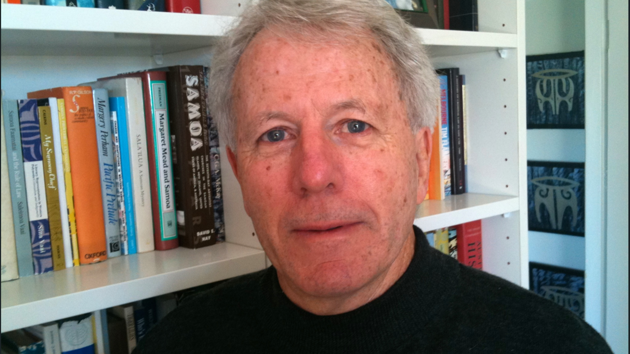 A portrait of Peter. He's standing in front of a wall of books. He is in a black sweater.