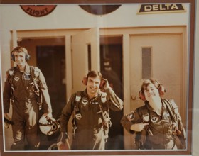 A picture showing three pilots in uniform walking away from a door. John is in the middle.