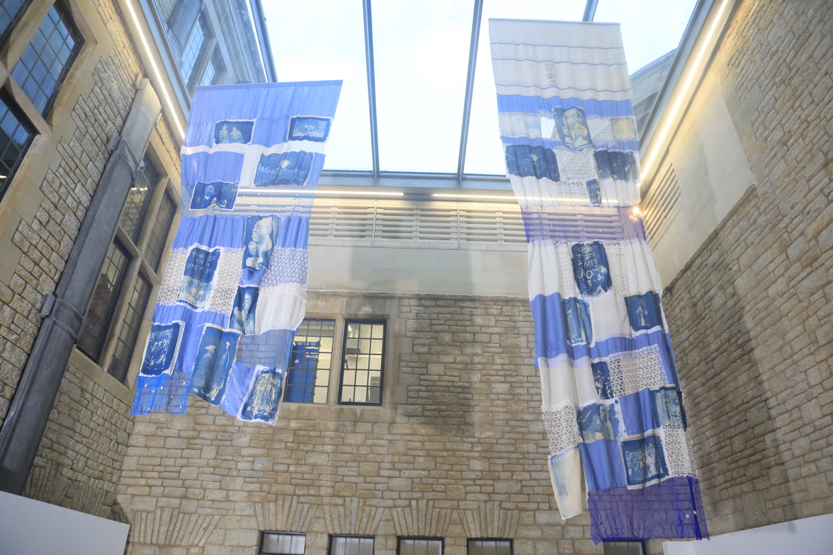 Two large blue patchwork textile pieces suspended from the glass ceiling.