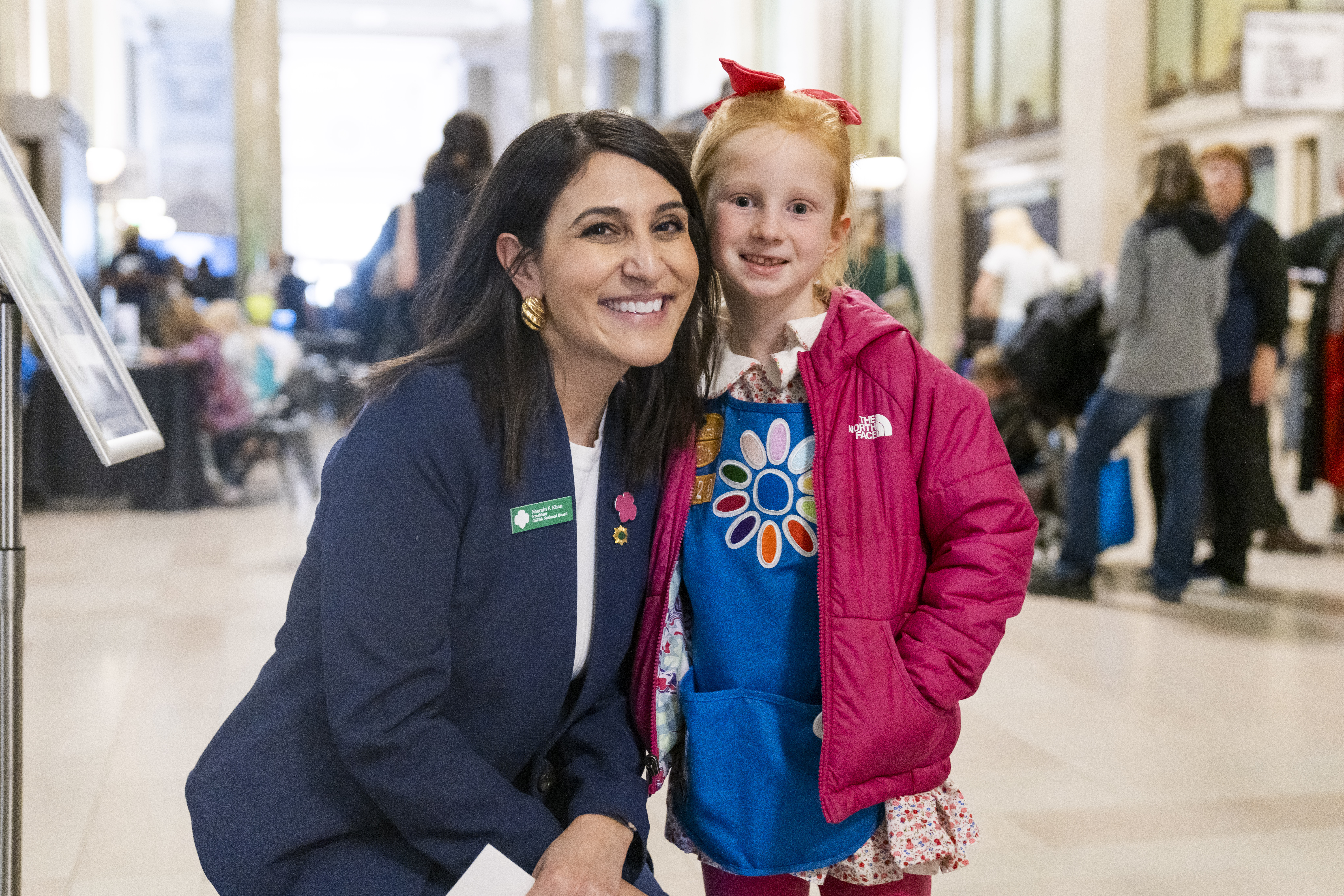 Noorain is bent down to be level height with a young girl who is wearing an aprol and pink jacket. They are both smiling, and stood in what appears to be a hall, with people milling around in the background.