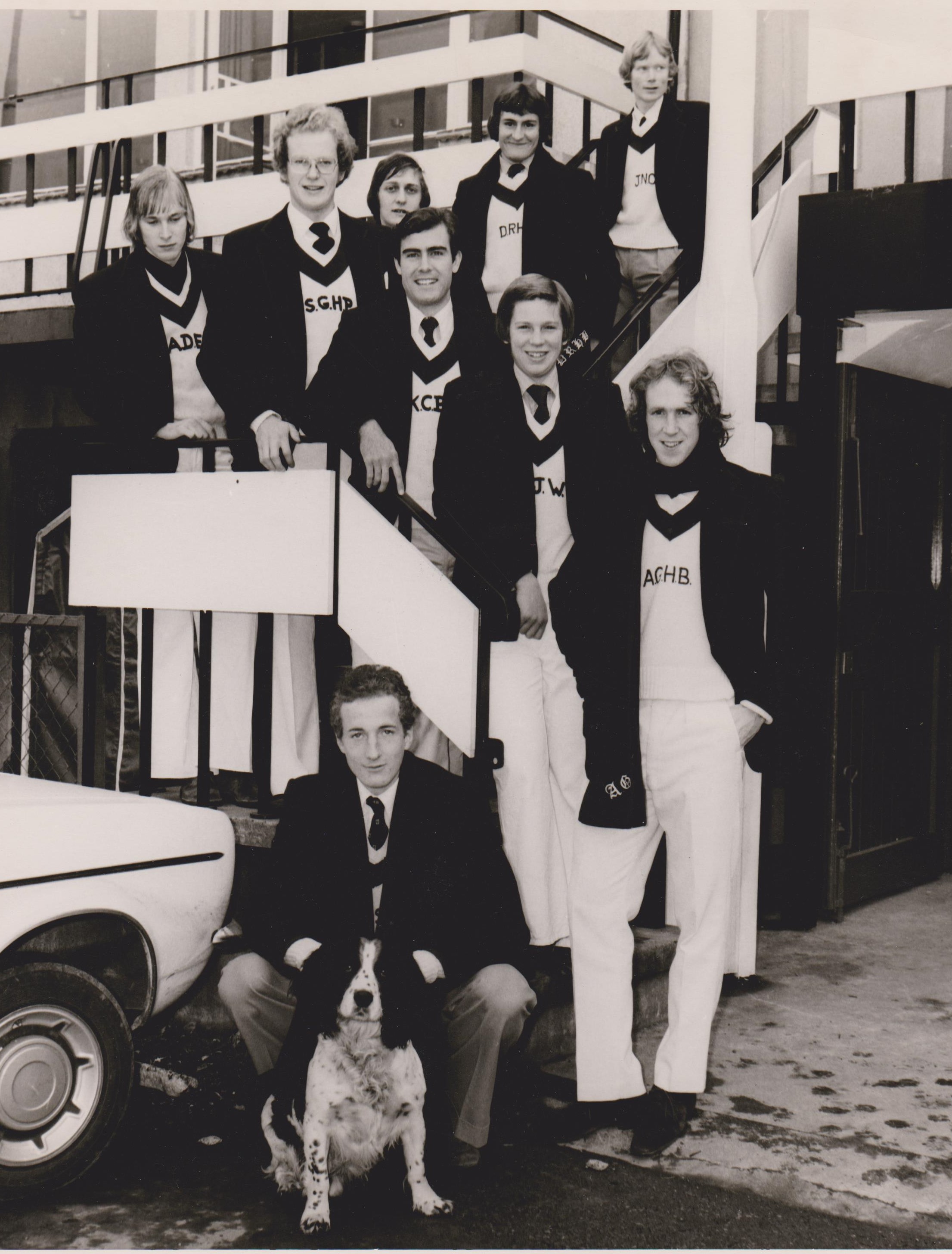 Black and white photo of a group of nine students posing around a staircase. Thre is a black and white dog photographed with them.