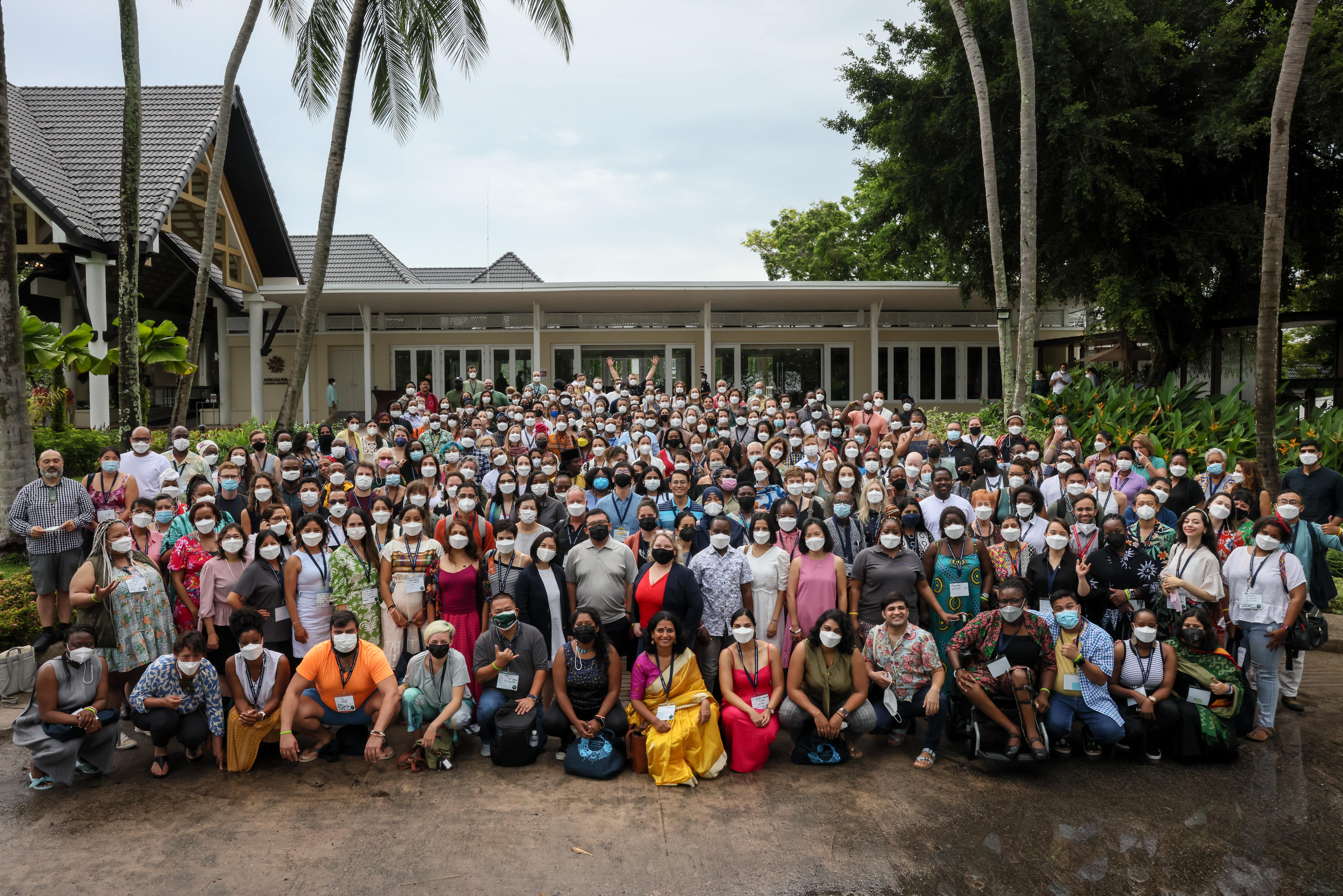 Image of a large gathering of over 100 people posing in front of a small white building surrounded by tropical plants.