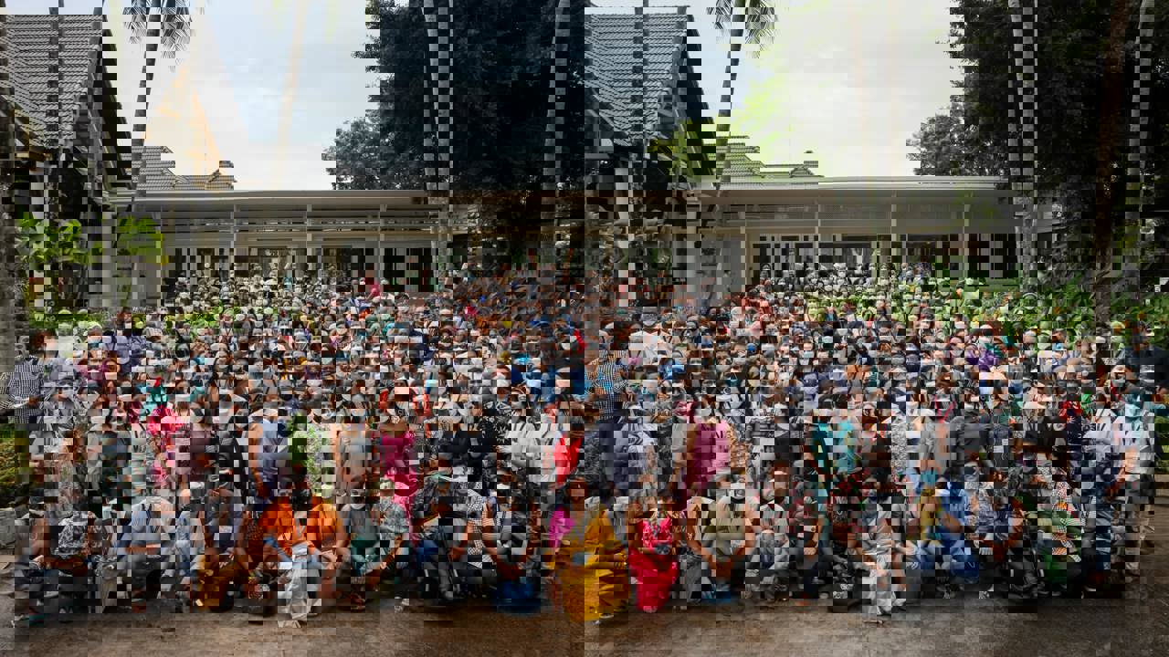 Image of a large gathering of over 100 people posing in front of a small white building surrounded by tropical plants.