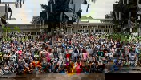 Image of a large gathering of over 100 people posing in front of a small white building surrounded by tropical plants.