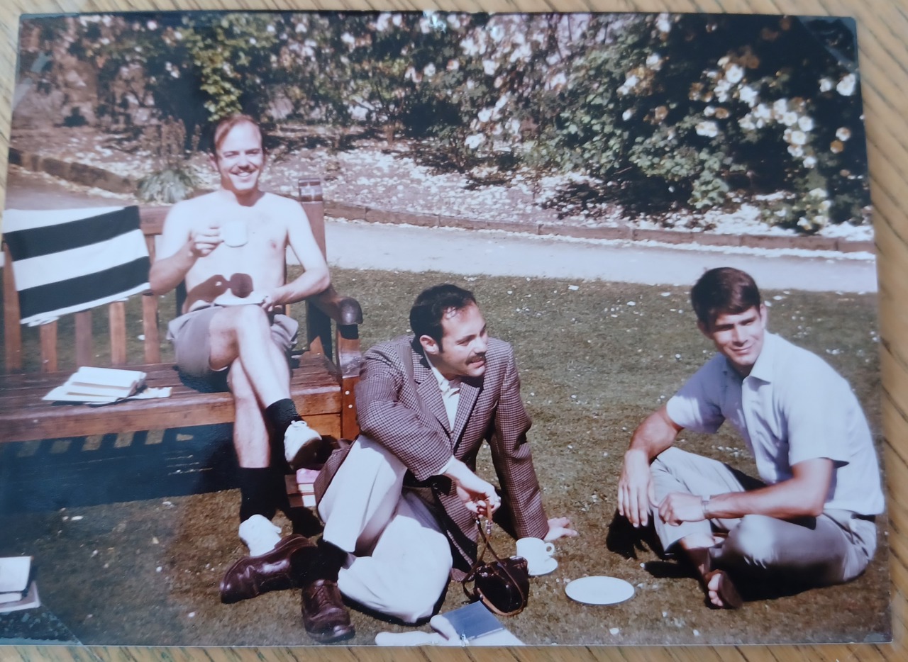 The picture shows a shirtless Goerge sitting on a bench with a cup in his hands. Eugene Ludwig is sitting on the grass near his feet and Steve Sturm is on his left. They both are dressed smartly.