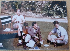 The picture shows a shirtless Goerge sitting on a bench with a cup in his hands. Eugene Ludwig is sitting on the grass near his feet and Steve Sturm is on his left. They both are dressed smartly.