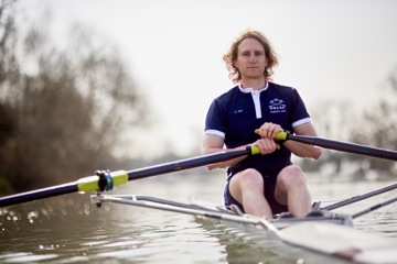 A Scholar rowing on the Isis on an overcast day. There are trees at the waters edge