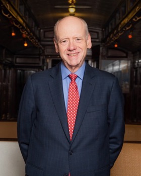 A headshot of Gary wearing a navy-blue suit and a red tie with white dots