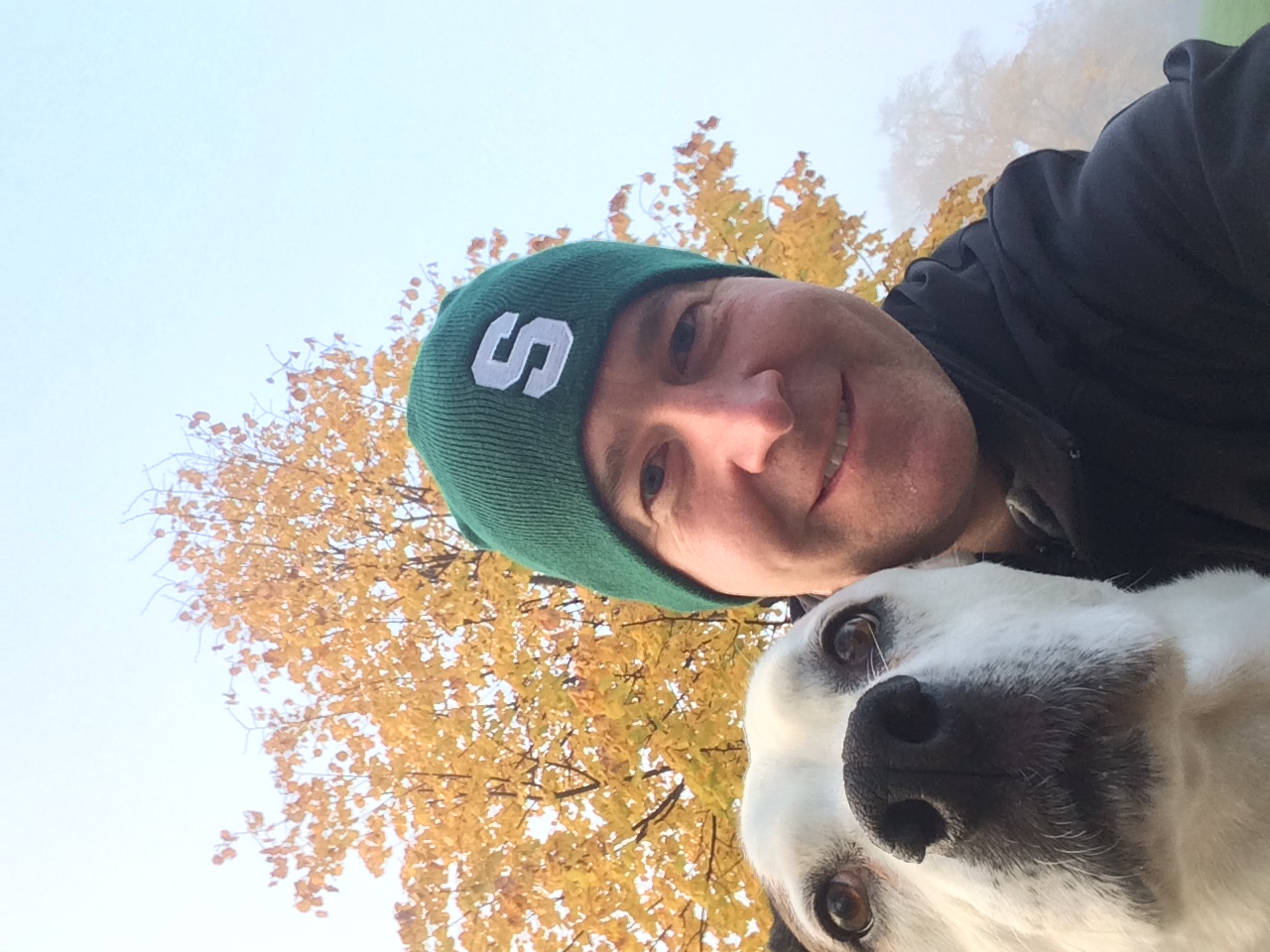 Close up Selfie of Dayne smiling in to the camera next to his white dog. There is a tree behind him