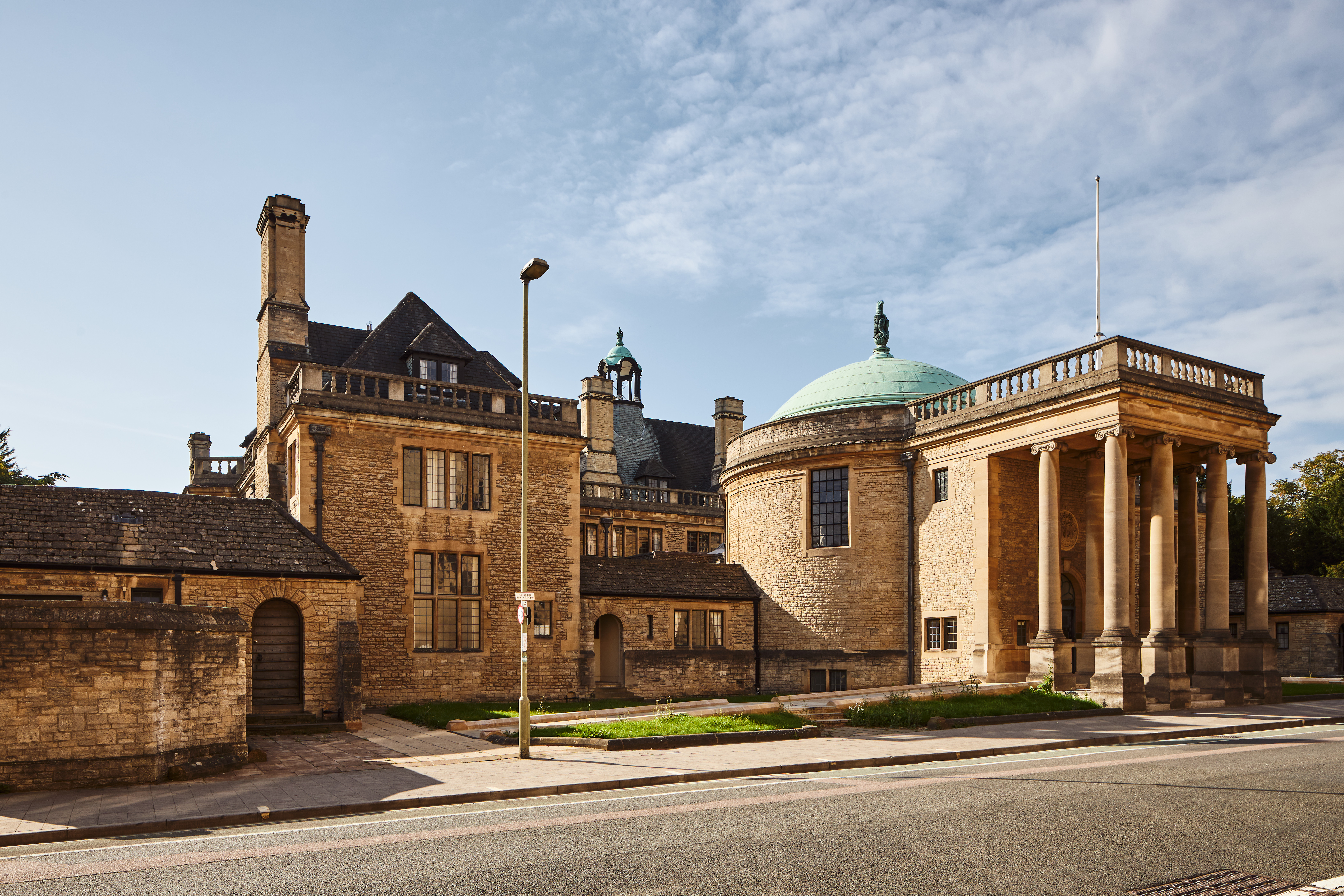 An image of Rhodes House from outside in the sun. It is a brown, stone building with a domed roof and pillars at the entrance.