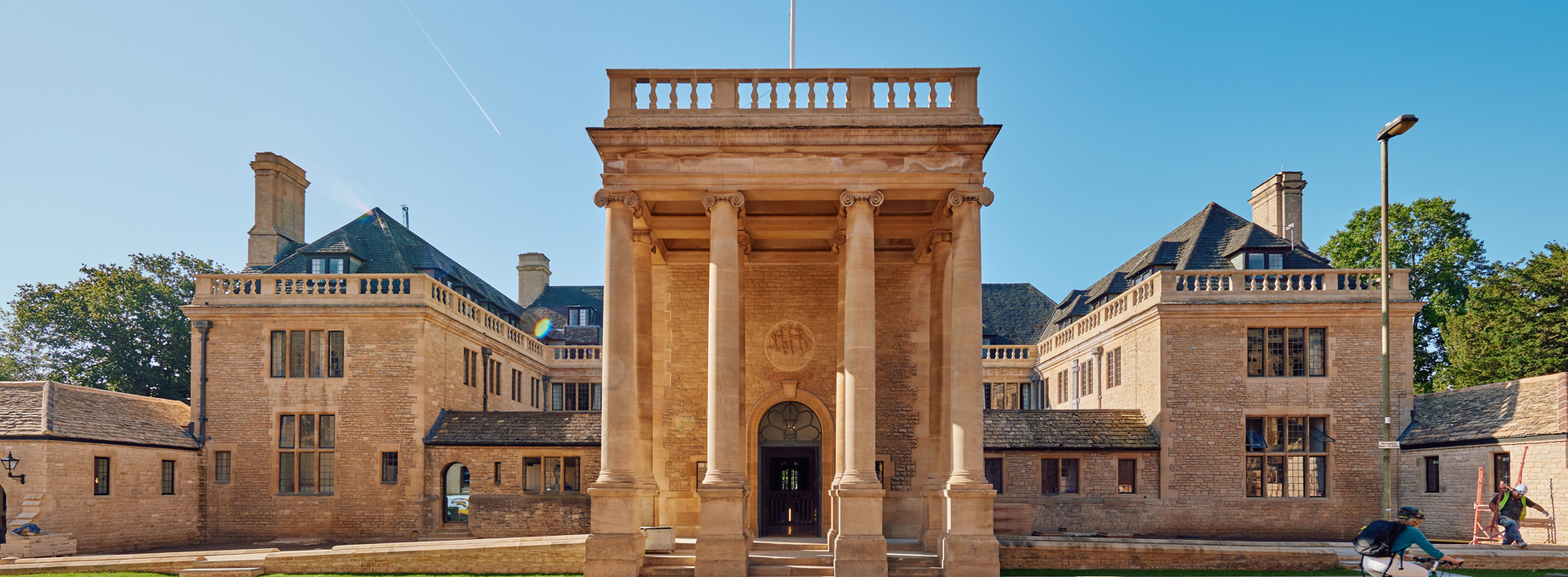 An Image of Rhodes House from the front. It is a stone building with four large pillars at the front and is illuminated by the sun under a clear blue sky.