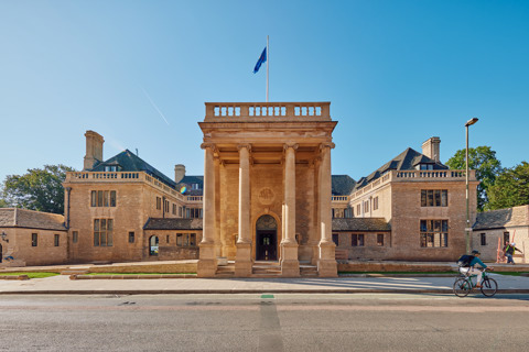 An Image of Rhodes House from the front. It is a stone building with four large pillars at the front and is illuminated by the sun under a clear blue sky.