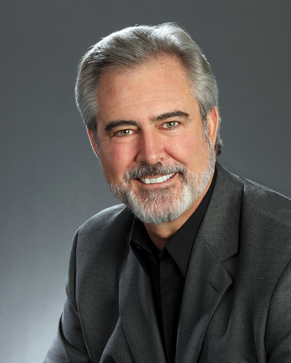 Headshot photo of Ken, wearing a black shirt, gray suit, smiling into camera.