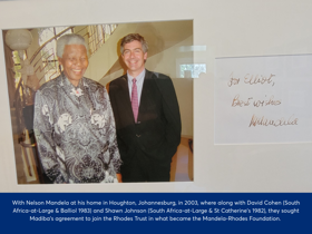 Photo of a framed image of Elliot standing to the right of Nelson Mandela, both smiling. The text to the right is a message from Nelson to Elliot saying 'best wishes'. Elliot was with Nelson Mandela at his home in Houghton, Johannesburg in 2003, where along with David Cohen (South Africa-at-Large & Balliol 1983) and Shawn Johnson (South Africa-at-Large & St Catherine's 1982), they sought Madiba's agreement to join the Rhodes Trust in what became the Mandela-Rhodes Foundation.